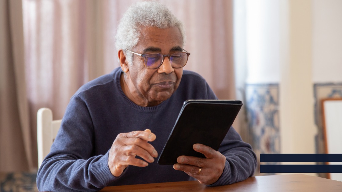 Älterer Mann mit Brille sitzt am Tisch und liest konzentriert auf einem Tablet