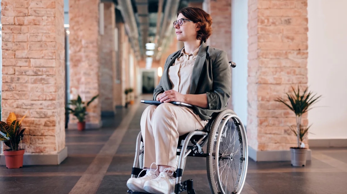 Junge Frau im Rollstuhl mit Tablet auf dem Schoß sitzt in einem modernen, hellen Gebäude und blickt optimistisch nach vorn