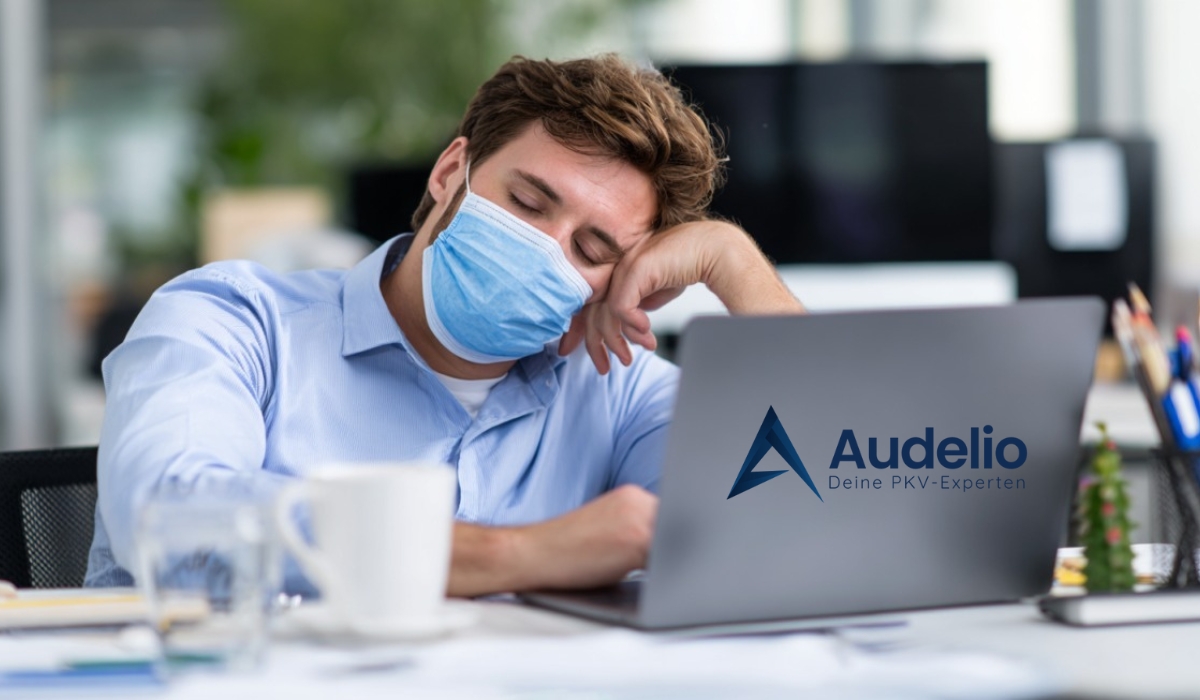 Ein Mann mit Gesichtsmaske sitzt erschöpft am Laptop im Büro, daneben das Logo von Audelio mit dem Slogan „Deine PKV-Experten“