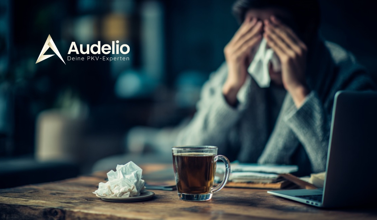 Eine Person sitzt krank mit Taschentuch am Tisch, vor ihr eine Tasse Tee und ein Laptop, daneben das Logo von Audelio mit dem Slogan „Deine PKV-Experten“